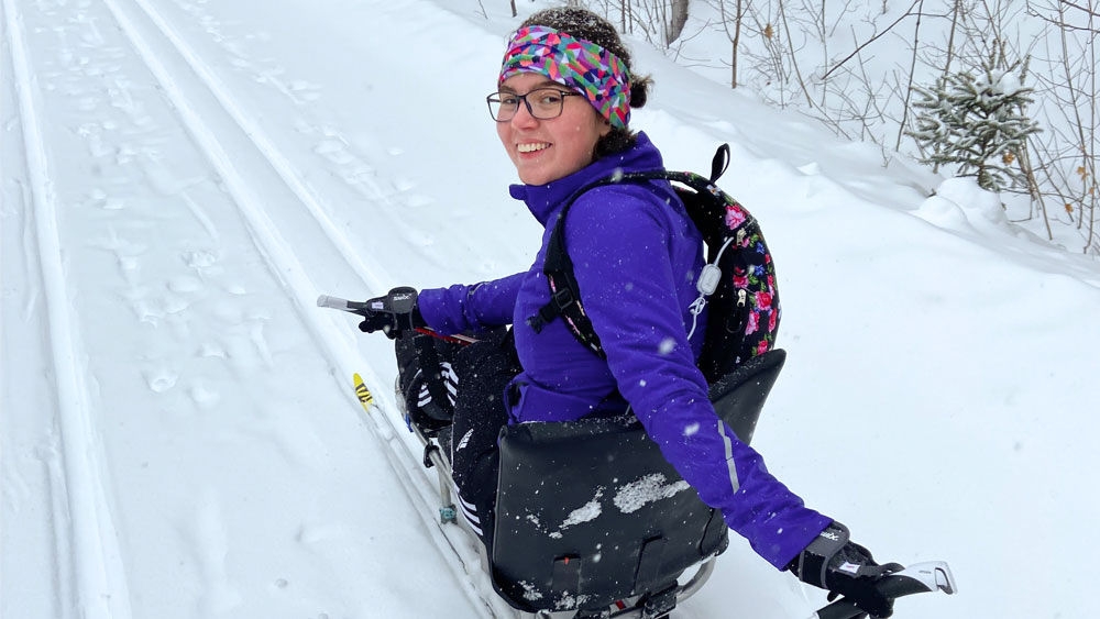 Una donna guarda la fotocamera con un sorriso sul viso mentre si siede sugli sci e scia per una strada piena di neve. 