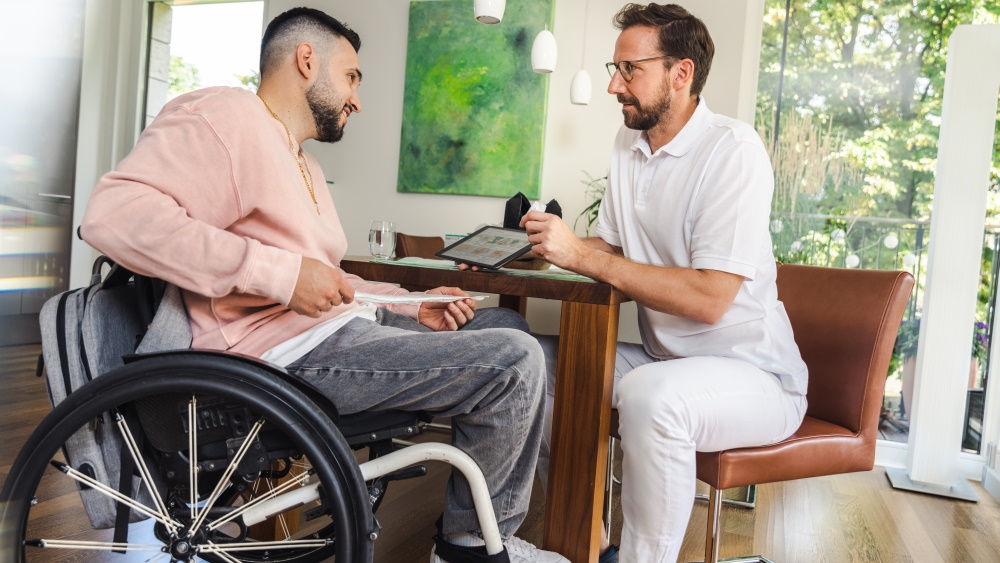 Un homme est assis dans un fauteuil roulant et tient un cathéter dans les mains. Devant lui, un professionnel de santé donne des instructions à l’aide d’une tablette