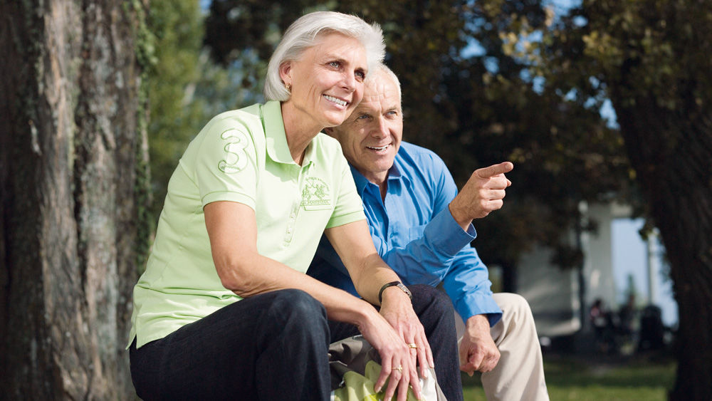 A middle-aged man and woman sit together in a forest, both smiling, a man pointing at something with his finger