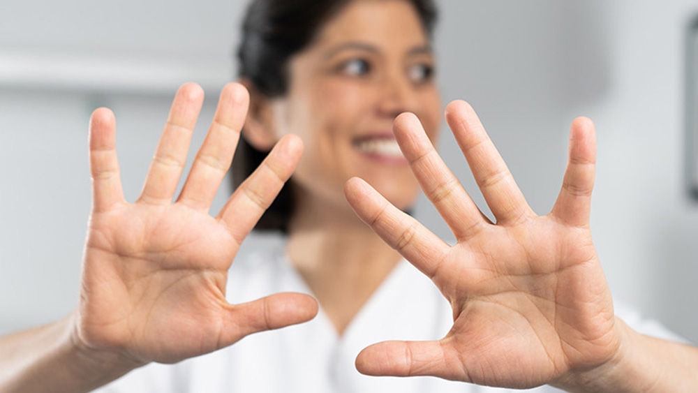 Eine Person trägt einen weißen Laborkittel und blickt in die Kamera. Beide Hände werden mit gespreizten Fingern nach vorne gestreckt. Das Gesicht der Person wird absichtlich verschwommen, um ihre Identität zu schützen. Im Hintergrund ist eine unscharfe Innenkulisse mit einem Bild an der Wand und einem Möbelstück, das wie ein Bett oder eine Couch aussieht.