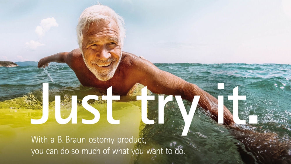 An elderly man with white hair is lying on a yellow surfboard, paddling in the ocean under a clear blue sky.
