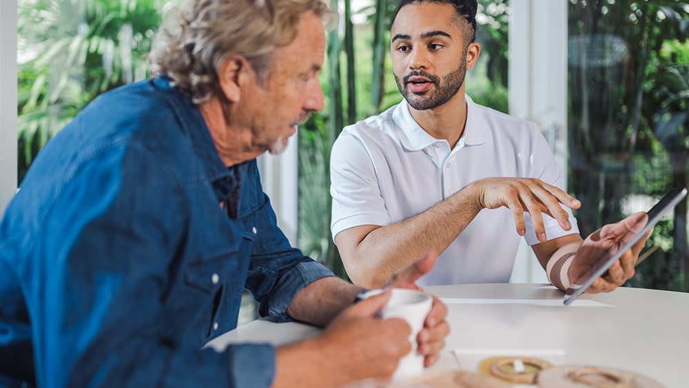 An elderly man sits at a table with a cup in front of him, listening to a man holding a tablet who is explaining something to him