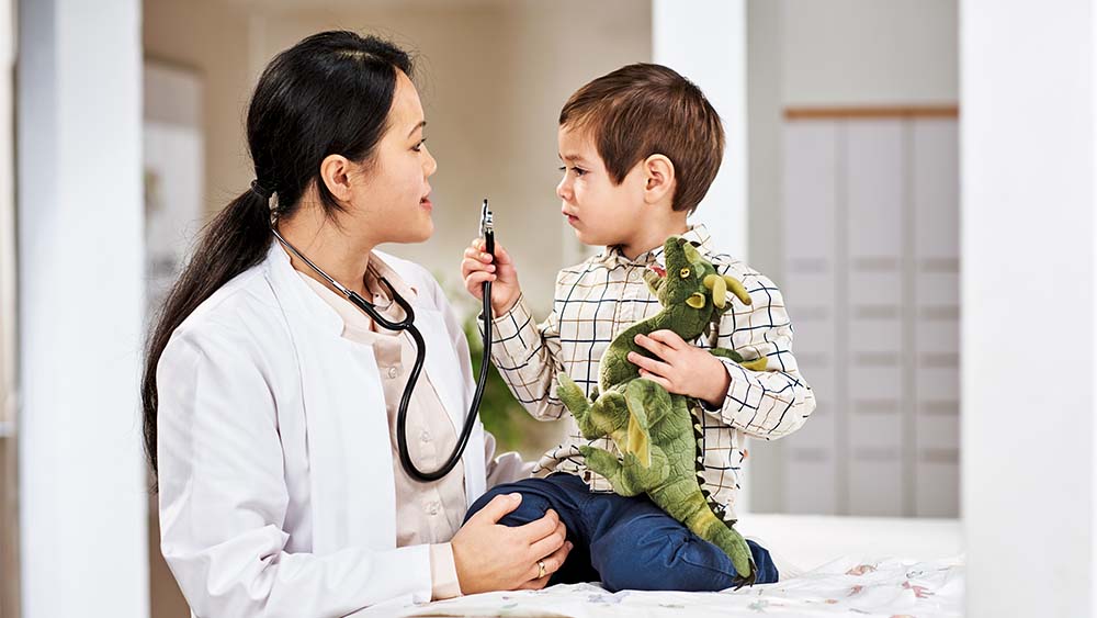 A female healthcare professional is talking to a small boy who is holding a toy dinosaur. The boy looks engaged and curious, while the healthcare professional has a warm and friendly expression