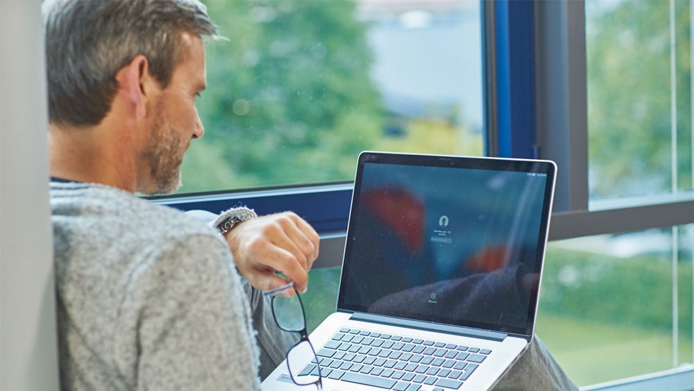 Eine sitzende Person hält einen Laptop auf dem Schoß. Mit der linken Hand tippen sie auf die Tastatur, während sie eine Brille in der rechten Hand halten. Auf dem Laptop-Bildschirm ist ein verschwommenes Kopfhörersymbol und die Aufschrift „Tippen, um zuzuhören“ zu sehen. Im Hintergrund ist durch ein großes Fenster eine grüne, natürliche Landschaft zu sehen.