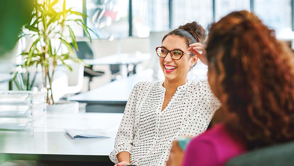 À droite, une femme est assise avec le dos de sa tête, à gauche, une femme est assise devant elle et regarde côté en riant. En arrière-plan, un bureau