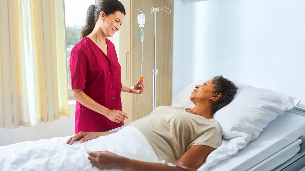Pflegekraft mit ecoflac plus auf einem Infusionsständer spricht mit einem Patienten, der im Bett liegt