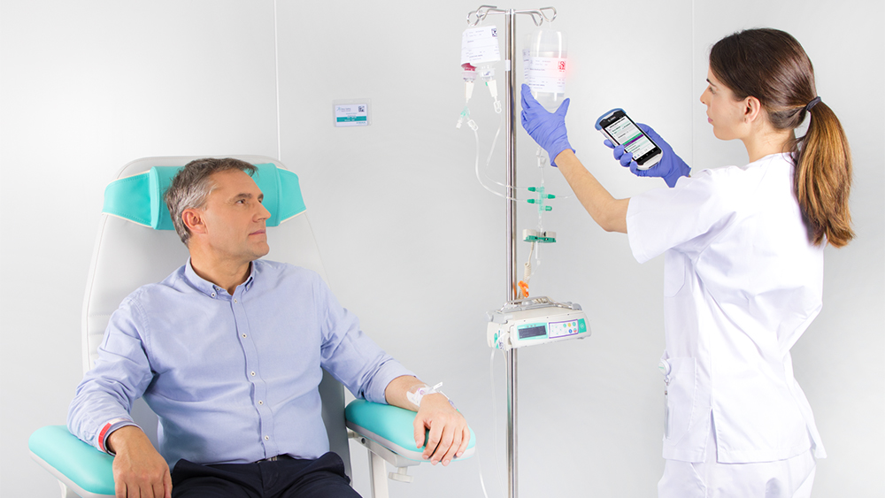 Billedet viser en patient, der sidder i en stol og modtager en infusion fra et medicinsk apparat fra B. Braun, mens en sygeplejerske indstiller infusionsdoseringen på apparatet.