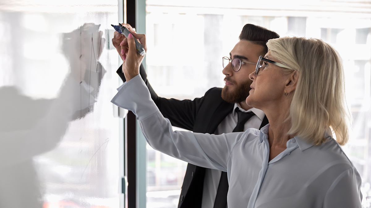 A man and a woman, dressed in business attire, writing ideas together on a whiteboard.