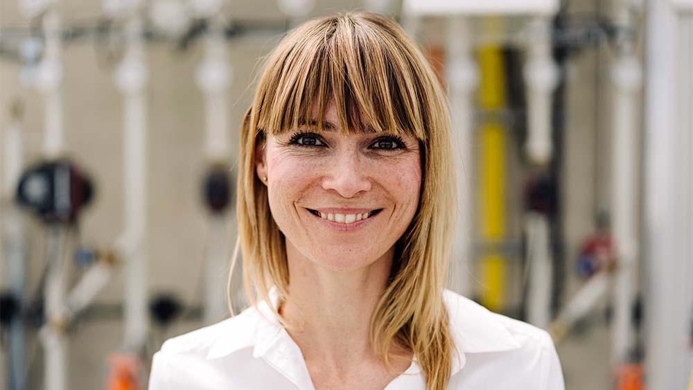 Gros plan sur une femme regardant l’appareil photo, souriant et portant une chemise blanche dans un environnement lumineux. 