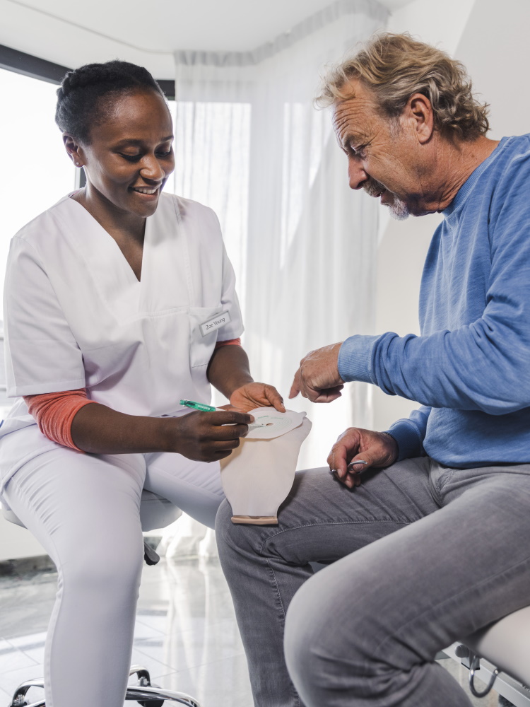 A female healthcare specialist is demonstrating with her hands how to use a stoma bag, while a man points at the bag with a questioning expression.