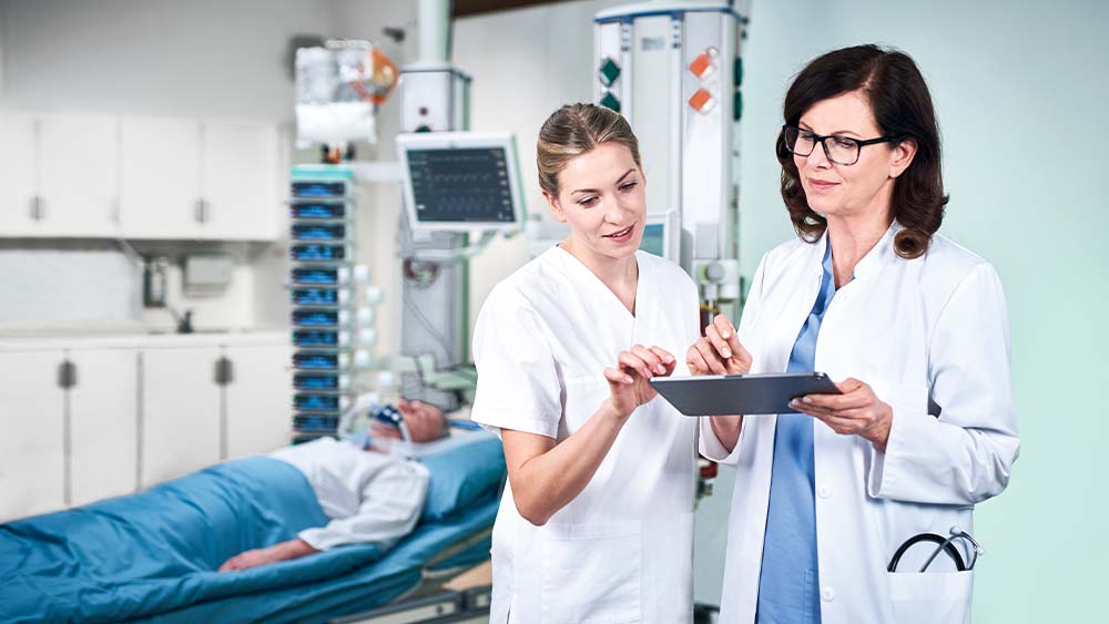 Lekarz i pielęgniarka omawiają dawkowanie inteligentnej infuzji B. Braun, która jest wprowadzana bezpośrednio z tabletu. Pacjenta na oddziale intensywnej terapii można zobaczyć w tle, połączony z wieżą pełną inteligentnych urządzeń infuzyjnych.