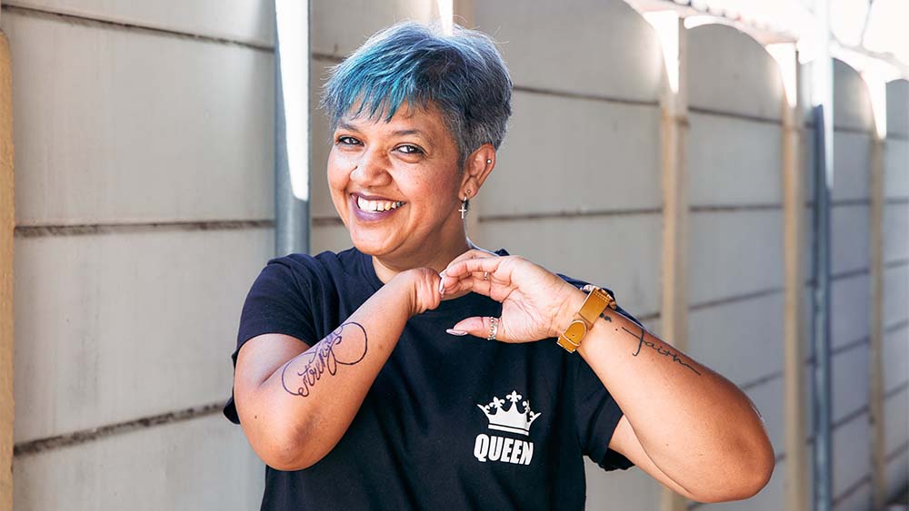 Uma mulher sem a mão direita, cabelos azuis e tatuagens está sorrindo, irradiando confiança e autoexpressão. 