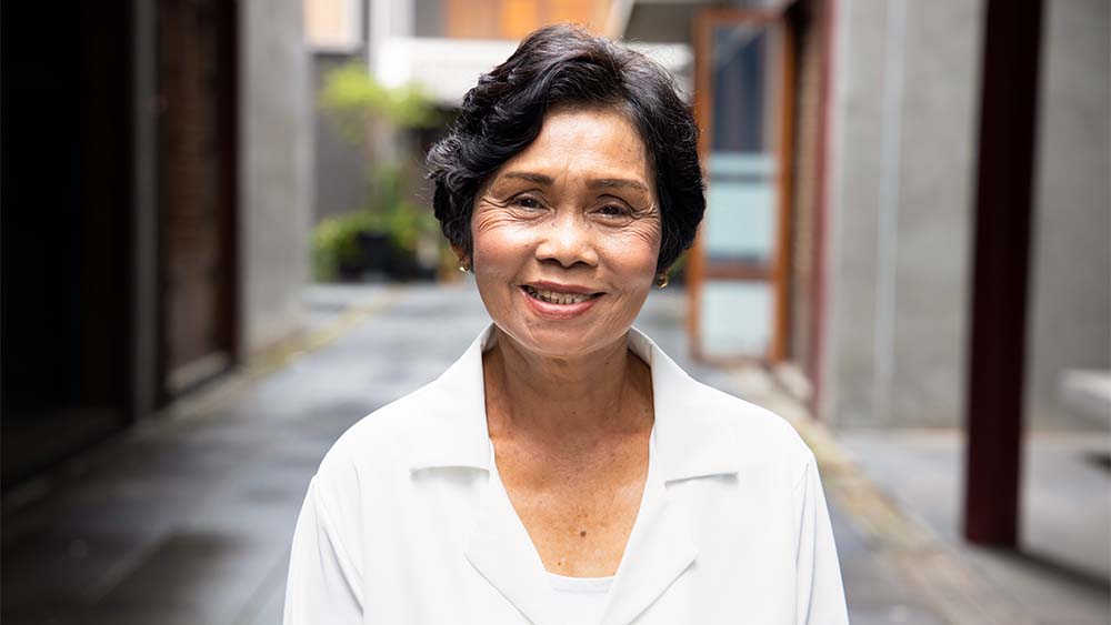 Uma mulher idosa com um sorriso caloroso, parada em um beco charmoso. 