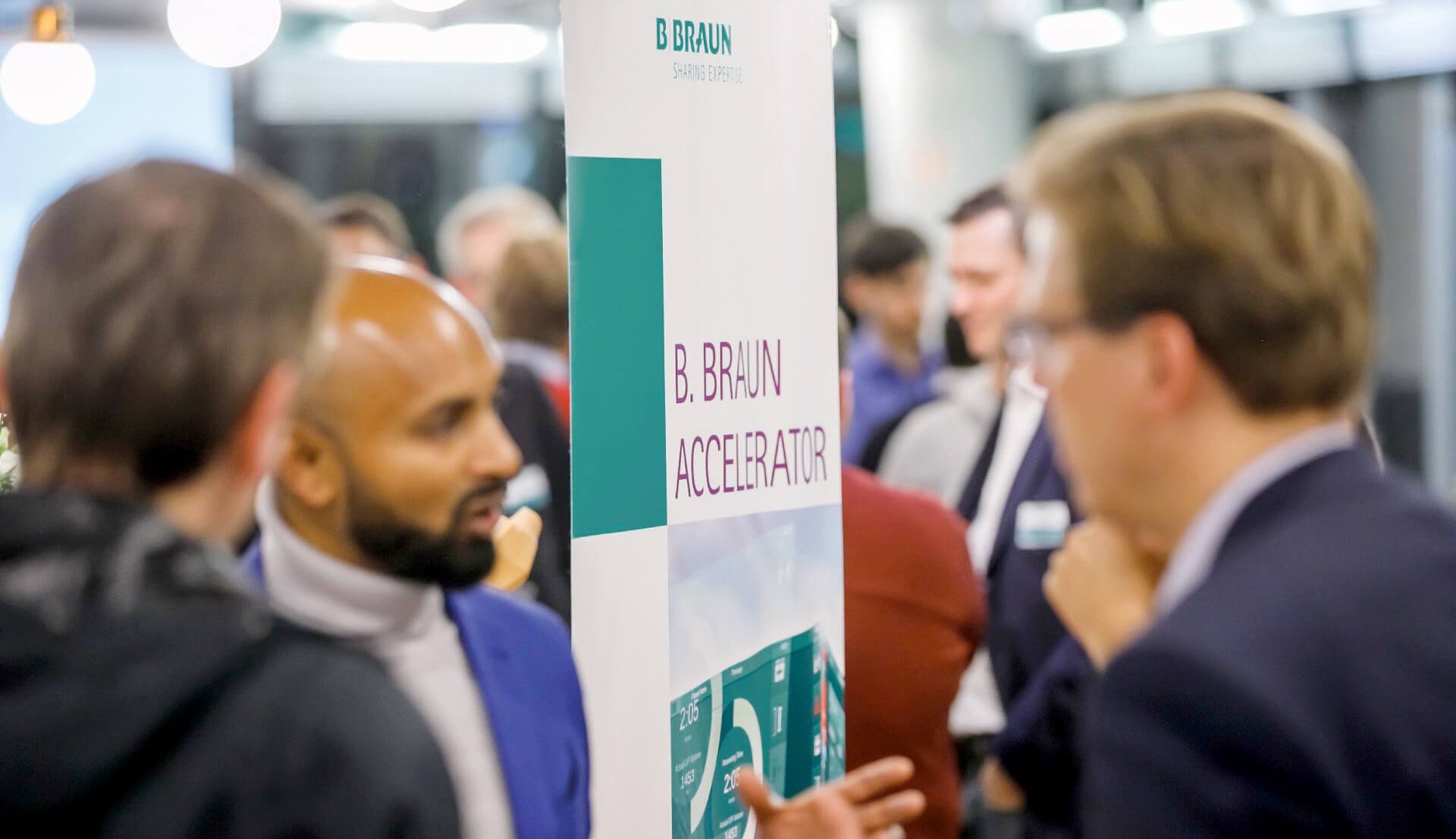 Một nhóm người thảo luận tại một hội nghị với một biểu ngữ về máy tăng tốc B Braun trong nền. 